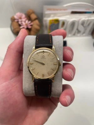 Klassisk Omega herrklocka i guld - Stilren vintage Omega klocka med rund guldfärgad boett och ljus urtavla. Klockan har ett brunt läderarmband med orange sömmar och minimalistiska indexmarkeringar. Perfekt accessoar för dig som gillar tidlös design och elegans.