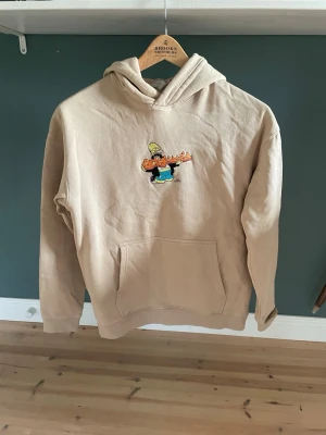 Beige hoodie från H&M Simpsons - Säljer en beige hoodie från H&M med tryck av Simpsons på bröstet. Hoodien har huva och en stor magficka framtill. Perfekt för dig som gillar streetstyle och vill ha något bekvämt och snyggt med en rolig detalj.