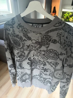 Grå mönstrad sweatshirt Versace Jeans couture  - Säljer en grå sweatshirt från Versace Jeans Couture med svart barockinspirerat mönster över hela tröjan. Tröjan har rund halsringning, ribbade muddar och är tillverkad i mjukt material. Perfekt för dig som gillar statement-plagg och exklusiv streetwear.