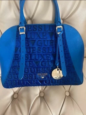 Blå Guess Luxe handväska  - Snygg blå handväska från Guess Luxe med präglad logotyp över hela väskan och silvrig metallbricka. Väskan har rundad form, perfekt för dig som vill sticka ut med en färgstark accessoar.