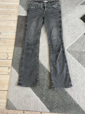 Grå bootcut jeans med låg midja - Snygga grå jeans med bootcut passform och låg midja. Klassisk femficksmodell med dragkedja och knapp framtill. Jeansen har en tvättad look och raka ben som släpper ut vid anklarna. Perfekta för en avslappnad och trendig stil.
