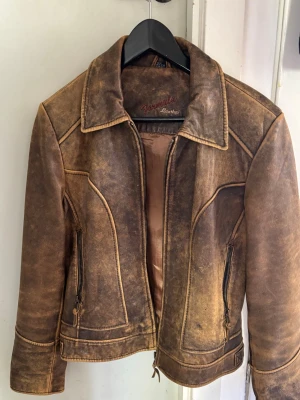 Brun vintage skinnjacka - Snygg brun skinnjacka från Giovanni Leather med vintage-look. Jackan har klassisk krage, dragkedja framtill och två fickor med dragkedja. Snygga sömdetaljer och justerbara spännen vid midjan ger extra stil.