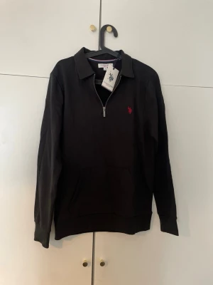 Svart halvzip från U.S. Polo Assn. - Svart långärmad tröja från U.S. Polo Assn. med halv dragkedja och klassisk krage. Tröjan har en röd broderad logga på bröstet och en praktisk magficka. Perfekt för en stilren och avslappnad look.