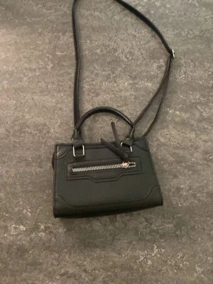 Svart liten handväska med axelrem - Snygg svart handväska i slät konstläder med både handtag och justerbar axelrem. Väskan har en silverfärgad dragkedja framtill och stilrena detaljer. Perfekt för dig som gillar minimalistisk och trendig design.