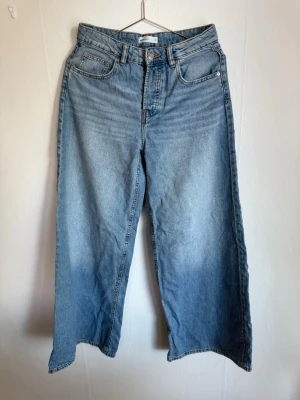 Vida ljusblå jeans med rå kant - Säljer ett par ljusblå jeans med bred passform och råa, ofållade benslut. Jeansen har klassisk femficksdesign och hög midja. Perfekta för en avslappnad och trendig look.