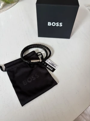 Svart flätat armband från BOSS - Stilrent svart armband från BOSS med flätad design i läder och en silverfärgad magnetlås med BOSS-logga. Levereras med original ask och tygpåse. Perfekt för dig som gillar minimalistiska accessoarer med premiumkänsla.