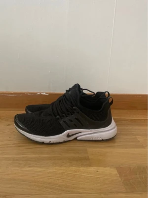 Nike Presto svarta sneakers - Svarta Nike Presto sneakers med vit sula och mesh-överdel. Skorna har en sportig design med elastiska snören och plastdetaljer vid sidorna. Perfekta för en avslappnad och trendig stil. Swoosh-logga på sidan och bekväm passform.