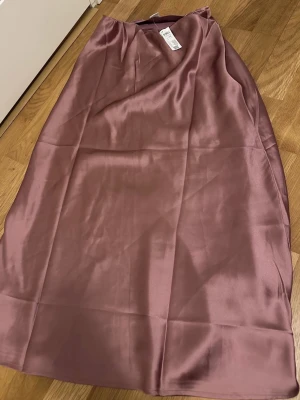 Lång rosa satinkjol från Lindex  - Säljer en lång, glansig lila kjol från Lindex i satinmaterial. Kjolen har en enkel och stilren design med lätt A-linjeformad siluett. Perfekt för dig som gillar en elegant och trendig look.