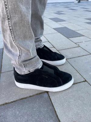 Arigato skor - Stilrena svarta sneakers från ARIGATO med ovandel i mocka och detaljer i textil. Skorna har en chunky vit sula och minimalistisk design utan synliga snören. Perfekta för dig som gillar en clean och modern look.