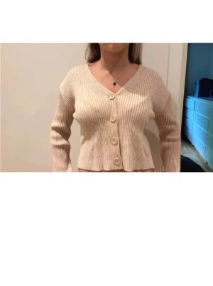 Beige ribbad kofta med knappar - Snygg beige ribbstickad kofta med v-ringning och stora knappar framtill. Modellen är croppad och har långa ärmar. Perfekt att slänga över en topp för en chill look.