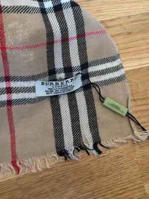 Beige rutig halsduk från Burberry - Klassisk beige halsduk från Burberry i 100% kashmir med det ikoniska rutmönstret i svart, vitt och rött. Halsduken har fransade kanter och är supermjuk och lyxig – perfekt för att lyfta vilken outfit som helst. Ett litet hål bara en