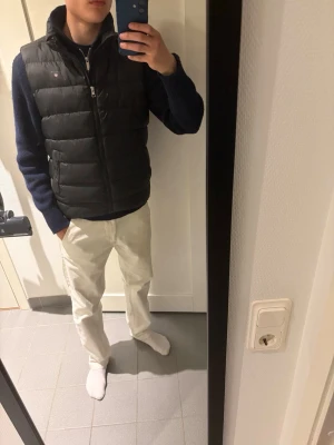 Svart dunväst från gant - Snygg svart dunväst från gant med diskret logga på bröstet. Västen har hög krage, dragkedja framtill och quiltad design. Perfekt att slänga över en hoodie eller tröja för extra värme och stil.