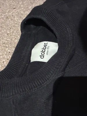 Svart sweatshirt från Dobber - En enkel svart sweatshirt från Dobber med rund hals och långa ärmar. Tröjan har ribbade muddar vid hals, ärmslut och nederkant. Perfekt för en avslappnad och stilren look.