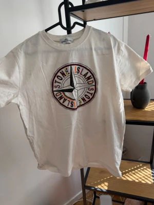 Vit Stone Island t-shirt med logga - Vit t-shirt från Stone Island med stor broderad logga på bröstet. Klassisk rund halsringning och korta ärmar. Perfekt för dig som gillar streetwear och vill sticka ut med en ikonisk design.