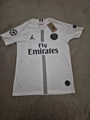 Vit PSG Neymar Jr matchtröja - Snygg vit PSG matchtröja med Neymar Jr och nummer 10 på ryggen. Tröjan har Fly Emirates-tryck, Jordan-logga och PSG-märke framtill samt Champions League-märke på ärmen. Tillverkad i lätt och ventilerande material, perfekt för fotbollsträning eller match. !Kan passa M!