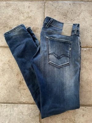 Replay blå slim fit jeans - Snygga blå slim fit jeans från Replay med klassisk femficksdesign och slitna detaljer. Jeansen har en cool tvätt med ljusare partier och kontrastsömmar. Bakfickan har Replay-logga och dekorativ söm. Perfekta för en trendig och avslappnad look.