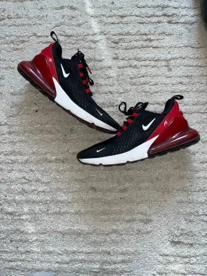 Nike Air Max 270 svart/röd sneakers - Nike Air Max 270 sneakers i svart och rött med vit sula och tydlig Air Max-dämpning i hälen. Ovandelen är i mesh för bra andningsförmåga och har röda detaljer vid snörningen. Swoosh-logga på sidan och Air 270-tryck på hälen.