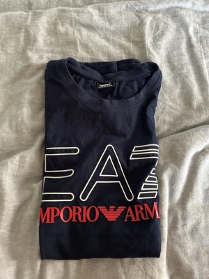 Mörkblå EA7 Emporio Armani t-shirt - Snygg mörkblå t-shirt från EA7 Emporio Armani med stort vitt och rött tryck på bröstet. Klassisk rund hals och korta ärmar. Perfekt för dig som gillar stilrena märkesplagg. Tillverkad i mjuk bomull för skön känsla.