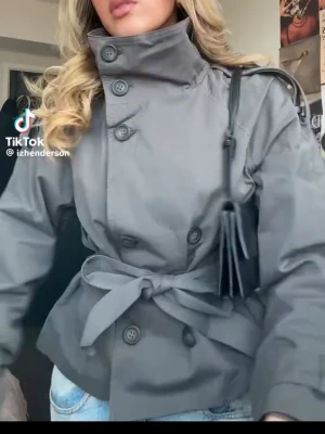 Mörkgrå trenchcoat med knytskärp från Bershka - Stilren mörkgrå trenchcoat med hög krage, knappar framtill och knytskärp i midjan från Bershka. Jackan har axelklaffar, långa ärmar med knäppning vid ärmslut och klassiska detaljer baktill. Perfekt för dig som gillar minimalistisk och tidlös stil.