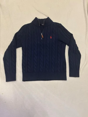 Mörkblå tröja Polo Ralph Lauren halv zip - Mörkblå kabelstickad tröja från Polo Ralph Lauren med halv dragkedja och klassisk röd logga på bröstet. Tröjan har hög krage, ribbstickade muddar och är tillverkad i mjukt material. Perfekt för lager-på-lager-stil. Den är för liten för mig så vill sälja skriv för mer frågor och om priset har använt den i några månader men den är i bra skick 