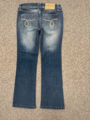Blå bootcut jeans från Streetwear - Snygga blå bootcut jeans från Streetwear Society Denim med slitningar framtill och bakfickor med gul dekorstickning. Klassisk femficksmodell och knappgylf. Perfekta för en avslappnad och trendig look.