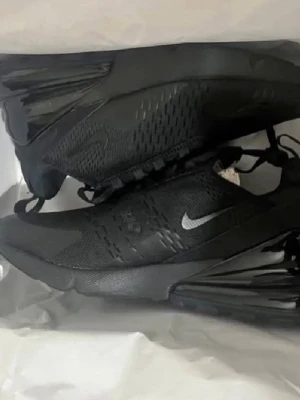 Nike Air Max 270 helsvarta sneakers - Snygga Nike Air Max 270 sneakers i helsvart med diskret silverfärgad swoosh. Skorna har en sportig siluett, mesh-ovandel för bra andningsförmåga och synlig Air Max-dämpning i hälen. Perfekta för en streetwear-look.
