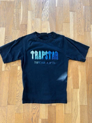 Svart Trapstar t-shirt med tryck - Svart t-shirt från Trapstar med färgglatt tryck på bröstet där det står 'TRAPSTAR' i blått och vitt. Klassisk passform med rund hals och korta ärmar. Perfekt för en streetwear-look.