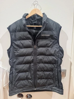Svart dunväst från Peak Performance - Snygg svart dunväst från Peak Performance med quiltad design och dragkedja framtill. Västen har hög krage och diskret logga broderad på bröstet. Perfekt för lager-på-lager och håller dig varm under kyliga dagar. Klassisk och stilren look.