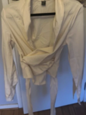 Krämvit omlottblus med långa ärmar - Stilren krämvit omlottblus från H&M med långa ärmar och knytband i midjan. Blusen har en djup v-ringning och är tillverkad i ett lätt, vävt material som ger en snygg och avslappnad look.