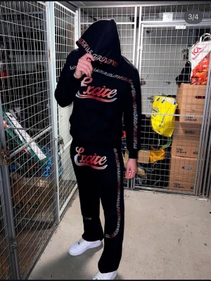 Svart hoodie från Excite med tryck - Svart hoodie från Excite med stort rött och vitt tryck på bröstet och ärmarna. Hoodien har huva och ribbade muddar. Snygg streetwear-stil med detaljer längs ärmarna och ett modernt snitt. Perfekt för dig som gillar en trendig och avslappnad look. Slut i lager 