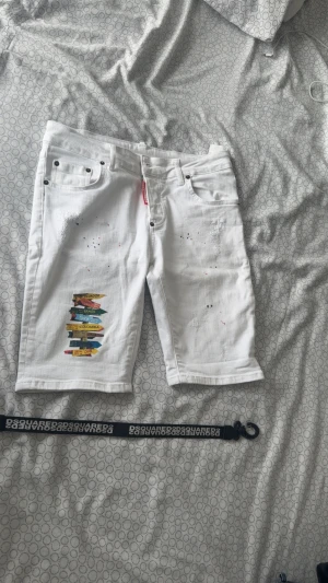 Vita shorts från Dsquared2 med tryck - Snygga vita shorts från Dsquared2 med färgstänk och coolt tryck med skyltar på ena benet. Klassisk femficksmodell med bälteshällor och knappgylf. Perfekta för dig som vill sticka ut med en unik och trendig stil.
