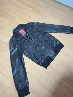 Jack & Jones svart skinnjacka – Strl XL (sitter som L) - Snygg svart skinnjacka från Jack & Jones Vintage Denim säljes. Klassisk modell med ribbade muddar och krage, flera fickor samt riktigt stilren passform. Perfekt till både vardag och kväll.  Storlek: XL Passform: Sitter mer som L Märke: Jack & Jones Färg: Svart Skick: Bra skick  Modellen på bilden är ca 185 cm lång och väger ca 78 kg för referens.