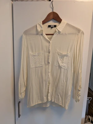 Vit skjorta från BikBok med fickor - Stilren vit skjorta från BikBok i ett tunt och mjukt material. Skjortan har två stora bröstfickor, klassisk krage och knäppning framtill. Perfekt för en avslappnad och trendig look.