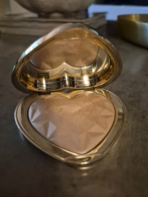 Hjärtformad highlighter i guld - Highlighter i en lyxig hjärtformad ask med spegel. Pudret har en skimrande champagnefärgad ton och en facettslipad yta. Perfekt för att ge glow till kindben och ansikte. Snygg och unik design som sticker ut i sminkväskan. Märke too faced, färg blinded by the light. Testad. Fin produkt, fint glow. Mer beige champagne glow! Lyxig förpackning!