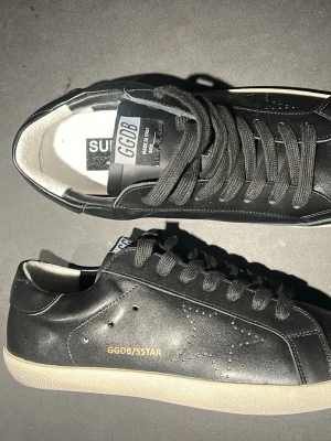 Svarta GGDB Superstar sneakers - Snygga svarta GGDB Superstar sneakers med klassisk låg profil och vit sula. Skorna har GGDB/STAR-tryck i guld på sidan, perforerad stjärndetalj och svarta skosnören. Tillverkade i läder med vit insida och GGDB-märke på plösen.