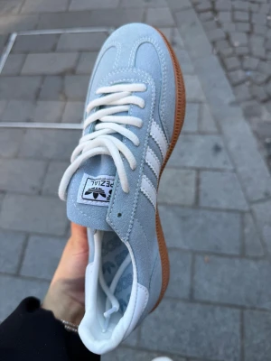 Adidas Spezial ljusblå sneakers - Säljer ett par Adidas Spezial sneakers i ljusblå mocka med vita detaljer och klassiska tre ränder på sidan. Skon har vit plös med Adidas-logga, vita skosnören och en brun gummisula. Guldfärgad text på sidan och retrovibe. Perfekt för dig som gillar stilrena och sportiga sneakers , finns i storlek 37-40.