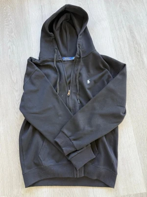 Svart hoodie från Polo Ralph Lauren - Klassisk svart hoodie från Polo Ralph Lauren med dragkedja, huva med dragsko och den ikoniska vita logon broderad på bröstet. Tillverkad i mjuk bomullsblandning och har ribbade muddar vid ärmar och nederkant. Perfekt för en chill och stilren look.