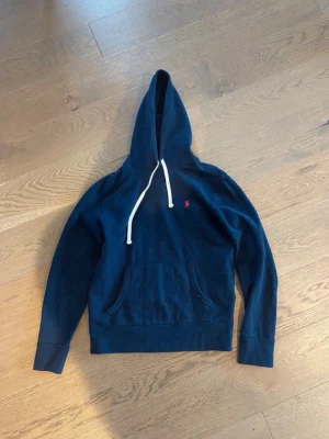 Mörkblå hoodie från Polo Ralph Lauren - Mörkblå hoodie från Polo Ralph Lauren med klassisk känguruficka framtill. Tillverkad i mjuk bomullsblandning, perfekt för en avslappnad och stilren look. Hör av dig vid frågor!!