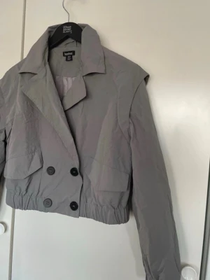 Grå cropped trenchcoat från Boohoo - Stilren grå cropped trenchcoat från Boohoo med dubbelknäppning och stora slag. Jackan har elastisk midja och långa ärmar. Tillverkad i ett lätt, tunt material som ger en modern look. Perfekt för dig som vill ha en trendig och avslappnad stil.