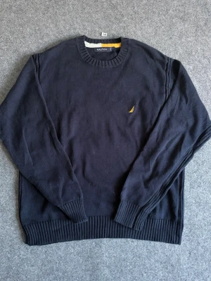 Mörkblå stickad tröja från Nautica - Mörkblå stickad tröja från Nautica med rund halsringning och ribbade muddar. Liten gul broderad logga på bröstet. Perfekt för dig som gillar klassisk och stilren design.