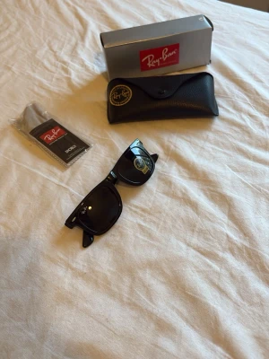 Svarta Wayfarer solglasögon Ray-Ban - Klassiska svarta Wayfarer solglasögon från Ray-Ban med ikonisk form och mörka glas. Kommer med originalfodral, putsduk och box. Perfekta för att lyfta din stil med en tidlös accessoar.