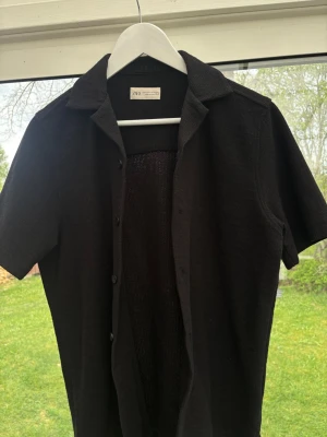 Svart kortärmad skjorta från Zara - Stilren svart skjorta från Zara med korta ärmar och klassisk krage. Skjortan har knappar framtill och är tillverkad i ett stickat material som ger en avslappnad men ändå trendig look. Perfekt att bära öppen över ett linne eller t-shirt.