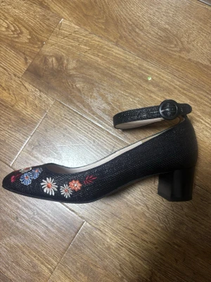 L.K. Bennett Klackskor med Blommor - Eleganta klackskor med ankelrem och en bekväm blockklack. Skorna har en spetsig tå och är prydda med färgglada broderade blommor på framsidan, vilket ger en charmig detalj. Ankelremmen är justerbar med ett spänne för en säker passform. Perfekta för både vardag och fest.  Skorna är små i storleken och passar bäst för dig som vanligtvis har storlek 38–38,5.