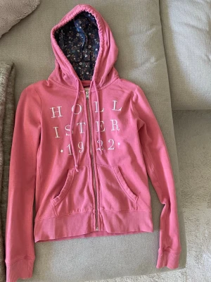 Rosa hoodie från Hollister  - Rosa zip-hoodie från Hollister med vit text 'Hollister 1922' på framsidan. Hoodien har en blommig innerfoder i huvan, två fickor och ribbade muddar. 