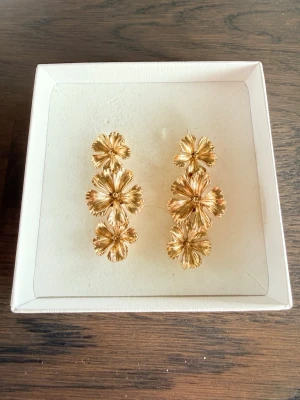 Guldiga blommiga örhängen - Säljer ett par snygga örhängen i guldmetall med tre hängande blommor på varje örhänge. De har en cool, detaljerad yta och ger en riktigt trendig look. Perfekta för dig som vill sticka ut med accessoarerna! Ej använda 💛