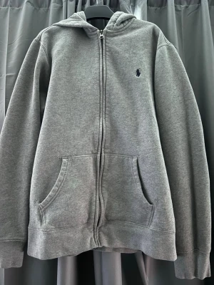 Grå zip-hoodie från Polo Ralph Lauren - En klassisk grå hoodie med dragkedja från Polo Ralph Lauren. Hoodien har huva, två stora fickor framtill och en diskret broderad logga på bröstet. Perfekt för chill dagar och enkel att matcha med jeans eller joggers. Dragkedjan är placerad på fel sida men när man stänger den så märks det inte