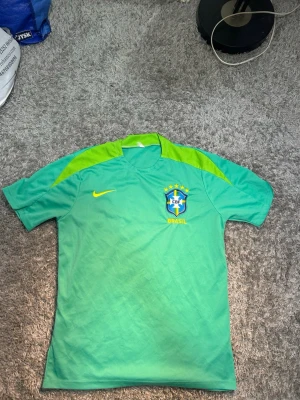 Grön Brasilien fotbollströja Nike - Snygg grön fotbollströja från Nike med Brasilien CBF-logga på bröstet och gula detaljer. Tröjan har korta ärmar och är tillverkad i ett lätt och ventilerande material, perfekt för fotbollsträning eller match.