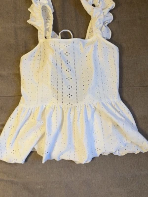 Vit volangtop med broderie anglaise - Söt vit topp med volangprydda axelband och broderie anglaise-mönster. Toppen har en luftig passform, öppen rygg med knytband och lätt utsvängd nederdel. Perfekt för sommarens alla tillfällen och ger en romantisk vibe. LITEN I STORLEK