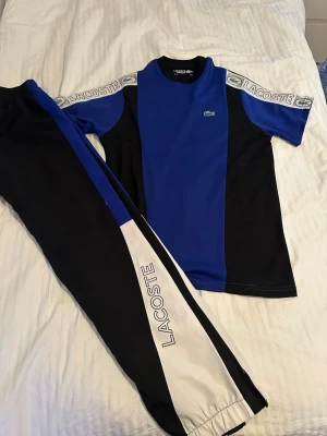 Blått och svart set från Lacoste - Snyggt tvådelat set från Lacoste med blå och svarta färgblock. T-shirten har korta ärmar, rund hals och logga på bröstet samt axelband med Lacoste-tryck. Byxorna är svarta med blå och vita detaljer samt Lacoste-tryck längs benet. Perfekt för en sportig look.