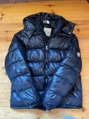 Svart dunjacka från Moncler - Snygg svart dunjacka från Moncler med glansig finish, huva och dragkedja framtill. Jackan har Moncler-logga på ärmen och praktiska fickor med dragkedja. Perfekt för kalla vinterdagar och riktigt trendig look.